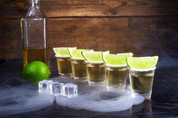 Mexican tequila gold in short glasses with salt, lime slices and ice on a wooden table. Smoke.