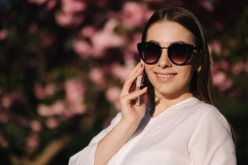 Portrait of attravtive young woman in white shirt and sunglasses use smartphone outside. Speel with someboby by the phone. Background of pink tree