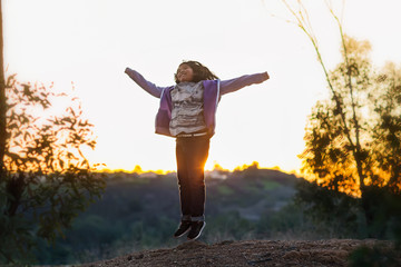 A young girl jumping with arms up on the top of a hill during a sunset.