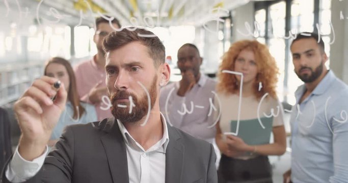 Bearded caucasian office worker writing formula on glass board and thinking . Group of male and female people standing and looking their chief near glass wall at corporate meeting.