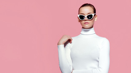 Fashion studio image of gorgeous elegant woman in white knitted golf and sunglasses posing over pink wall.