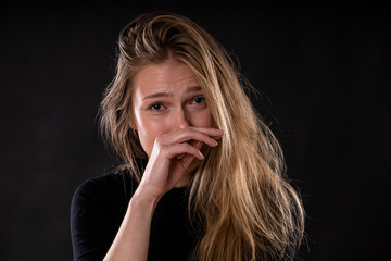 young beautiful blonde girl with a sad expression and tearful eyes wipes tears with her hand on a dark background