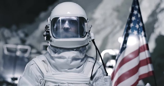 NASA spaceman in special white armor with a helmet standing in profile to the camera and then turning face to the camera and smiling with a USA flag on the background. Close up. Portrait on the Moon.
