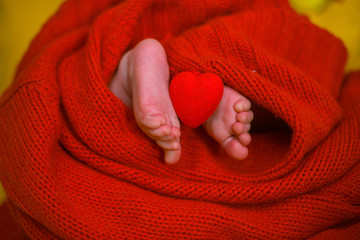 Red heart on baby legs. The legs of the newborn on a red background. A baby wrapped in a red blouse. Valentine's Day