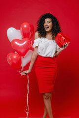 happy black woman with heart shaped gift box and colorful balloons isolated on red background