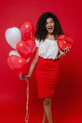 happy black woman with heart shaped gift box and colorful balloons isolated on red background