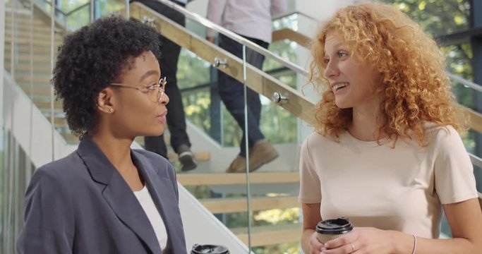 Two diverse race women office workers standing in office corridor and talking. Close up of young successful female coworkers drinking cofee, having pleasent talk and laughing.
