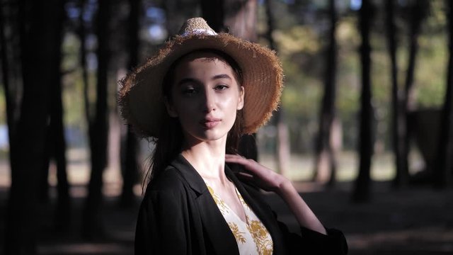 lady in straw hat and black jacket poses standing in forest under summer sunlight against blurry trees slow motion closeup