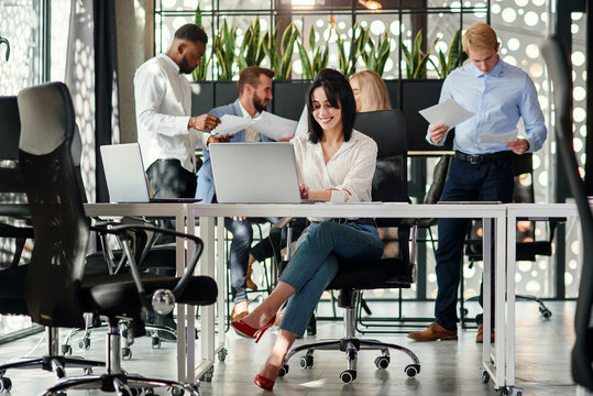 Pretty female office manager sits at her working table and uses two laptops in office on the background of her office colleagues