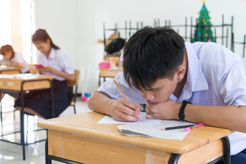 Test or Exam in school concept: Asian boy Students thinking reading paperwork at examination room with smiling,sitting learning lessons on wood desk chair doing final testing in classroom with uniform