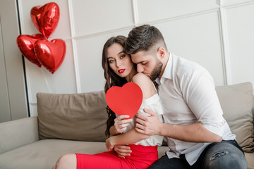 man and woman with heart shaped valentine card and red balloons sitting on couch at home