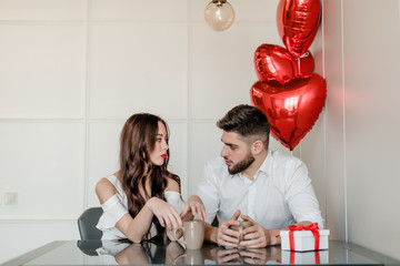couple man and woman drinking from cups in the morning with present in gift box and heart shaped balloons at home