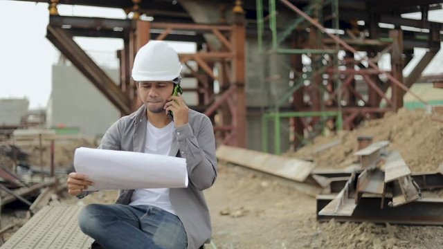 Asian construction engineer worker with protective helmet Talking on the phone and Check blueprints paper at building construction site