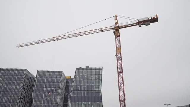 Rotating crane on construction site with office building in background, low angle wide shot
