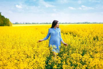 Fototapeta premium Beautiful pregnant girl walking on a rape field, stylish expectant mother in a blue dress