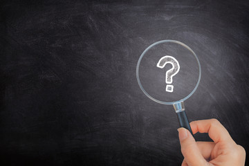 Business Concept : Female hand holding magnifying glass and looking at Question Mark icon symbols on black chalkboard.