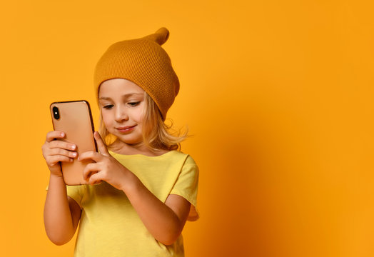 Little blonde kid dressed in t-shirt and hat, posing with smartphone against yellow background. Technology, children, internet. Close-up shot