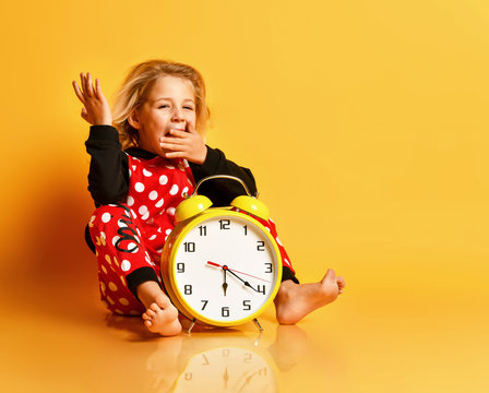 Little blond girl red dotted home clothing sitting on floor with big yellow alarm clock and yawning over yellow background