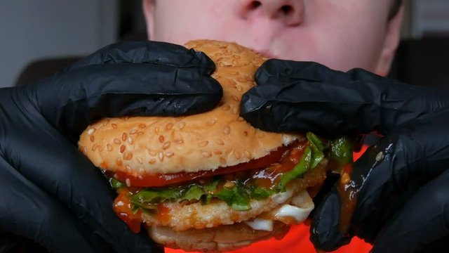A teenager eats a big juicy hamburger with an egg, a tomato cutlet, salad and red sauce. Hands in special black gloves hold junk food, fast food. Face and mouth close up view.