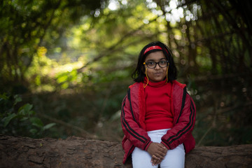 A cheerful Indian Bengali brunette baby girl with red woolen sweater sitting on a wooden log within a forest in a winter morning in green background. Girl in the park and Indian lifestyle, winter.
