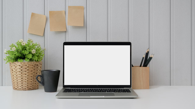 Close up view of workspace with blank screen laptop, stationery, sticky note, coffee cup and tree pot on white table with plank wall