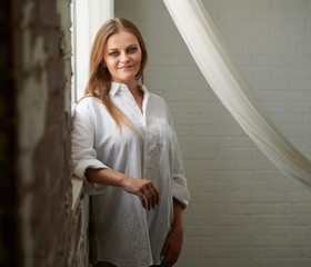 Fototapeta premium Stunning young Caucasian woman poses in studio wearing a men's white button-up shirt - back to window