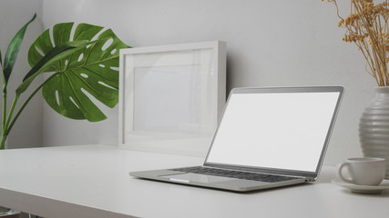 Close up view of workspace with blank screen laptop, mock up frame and ceramic vase on withe desk with white wall