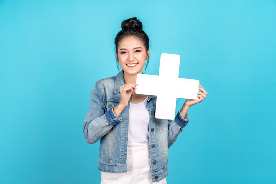 Happy asian woman standing and holding plus or add sign on blue background. Cute asia girl smiling wearing casual jeans shirt and showing join sign for increse, upgrade and more benefit concept