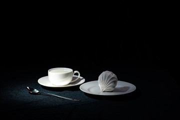 A saucer with a coffee Cup filled with milk and a plate with marshmallows on a black cloth background