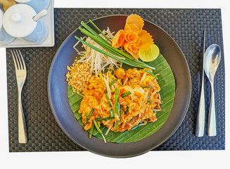 Pad Thai Shrimp on a banana leaf plate placed on a black plate. With knives, spoons, forks