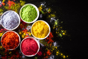 Holi festival celebration. Traditional Indian Holi colours powder decoration with paints. Top view of Organic Gulal colors in bowls, spices, rustic on black background for Holi festival.
