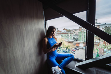 Pretty girl sitting on the window sill with smartphone in hands.