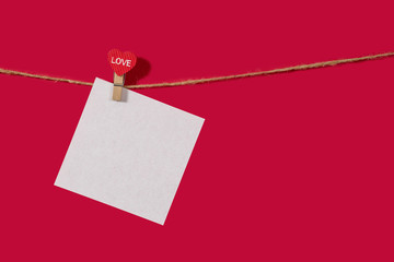 A white piece of paper on rope with a clothespin, with space for text. Mockup notes on a red classic background. Concept of recognition of love, wedding, romantic relationship, Valentine's day