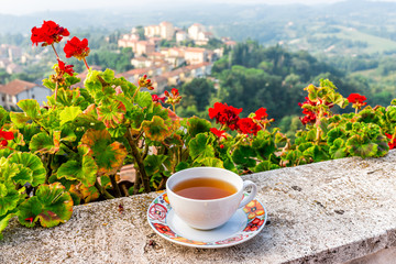 One black tea cup on balcony terrace railing by red geranium flowers outside in Italy with mountain...