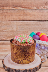 Easter composition. Easter cake on a stand made of a slice wood and a white basket on wheels with colorful Easter eggs on a wooden background. With copy space