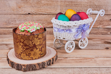 Easter composition. Easter cake on a stand made of a slice wood and a white basket on wheels with colorful Easter eggs on a wooden background. Close-up