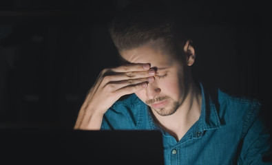 Tired young man keeping eyes closed and touching forehead while working at computer late, isolated black background