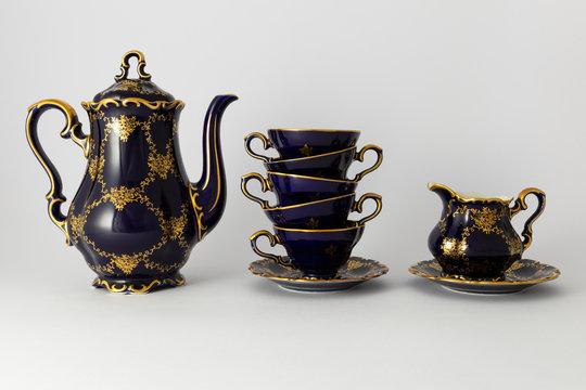 Closeup of a beautiful cobalt blue colored vintage porcelain tea set with golden floral pattern on white background. The set includes a tea pot, a milk jug and a stack of tea cups.
