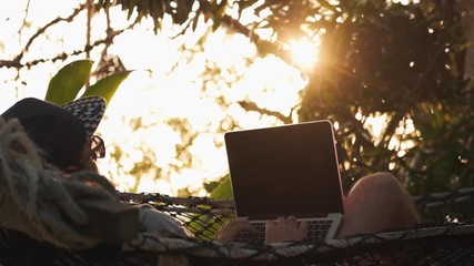 Man working with laptop relaxing in a hammock on sunset background. Digital nomad concept, remote worker, location freelance entrepreneur.