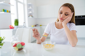 Photo of pretty upset housewife holding spoon don't want eat milk breakfast cornflakes tired of dieting bored sitting table white light kitchen indoors