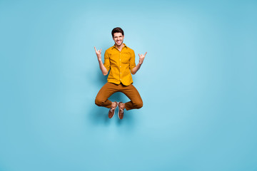 Full length photo of funny guy jumping high raising hands showing horns rejoicing rock concert wear yellow shirt pants isolated blue color background