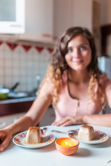 Obraz na płótnie Canvas Sweet panna cotta dessert food with bokeh of young happy woman smiling sitting at kitchen table looking at camera indoors in Italy romantic date