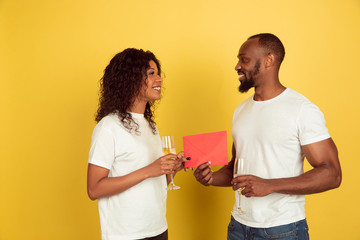 Giving red envelope. Valentine's day celebration, happy african-american couple isolated on yellow studio background. Concept of human emotions, facial expression, love, relations, romantic holidays.