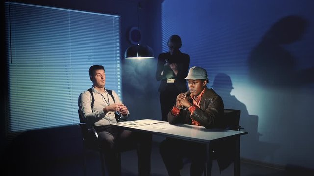 Black prisoner in handcuffs having interview with caucasian detective in interrogation room. Female assistant making notes of the interrogation.