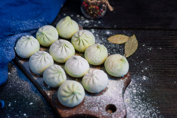 Frozen khinkali on a wooden board, sprinkled with flour. In the background pepper and bay leaf. On a black wooden background.