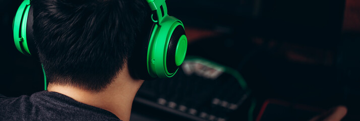 banner photo. Back view of gamer in green professional headphones using computer for playing game.