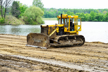 Yellow crawler tractor clears the construction site for recreation on the lake. - obrazy, fototapety, plakaty