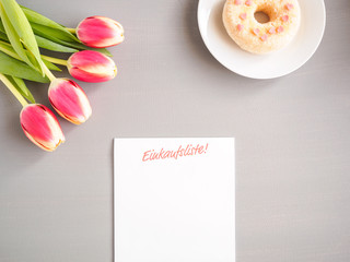 Unbeschriebener Einkaufszettel mit rosa Tulpen und einem Donat auf einem grauen Tisch, Draufsicht, Notiz