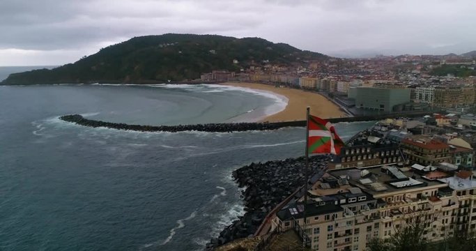 Aerial reveal of coastal San Sebastian, Spain with Basque flag in foreground.