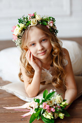 Kid - girl with blond hair in a flower crown holds in his hands a bouquet of fresh flowers. Beautiful little girl in a bright studio lying on the floor and looks into the camera